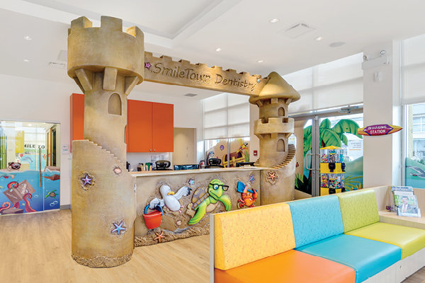 A brightly lit children's dental office features a sand castle-themed reception desk decorated with sea creatures. Colorful, cushioned benches line the waiting area, and whimsical beach murals decorate the walls.