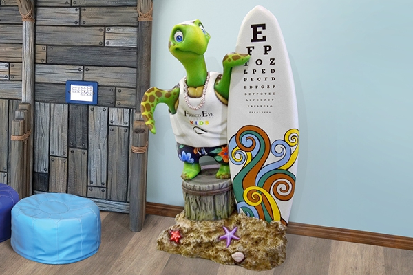 A cartoon turtle statue wearing a shirt with the business logo stands next to a surfboard with an eye chart printed on it, creating a perfect photo op in a colorful optometrist office filled with beach-themed decor.