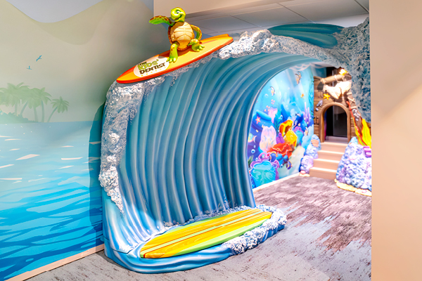 A colorful indoor display featuring a large, sculpted blue ocean wave with a yellow surfboard and a cartoon turtle on top. The wave leads the way to a sea-themed children's paly area with murals and a beach scene.