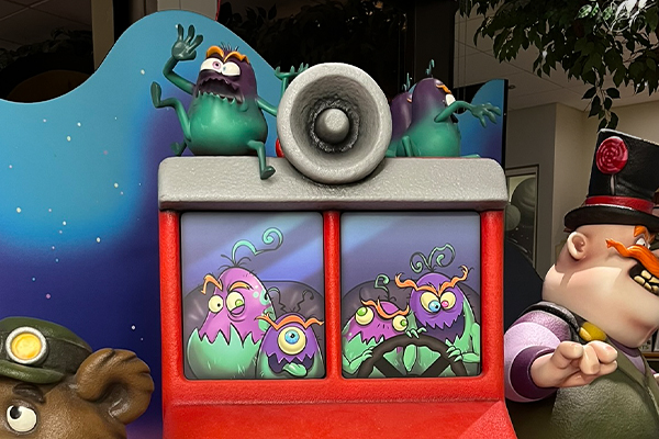Colorful cartoon tooth bugs ride a red bus with a loudspeaker on top. Some sugar bugs peek out the windows, while one on the roof waves excitedly. Cavitar, a man in a top hat stands on the right side of the image.