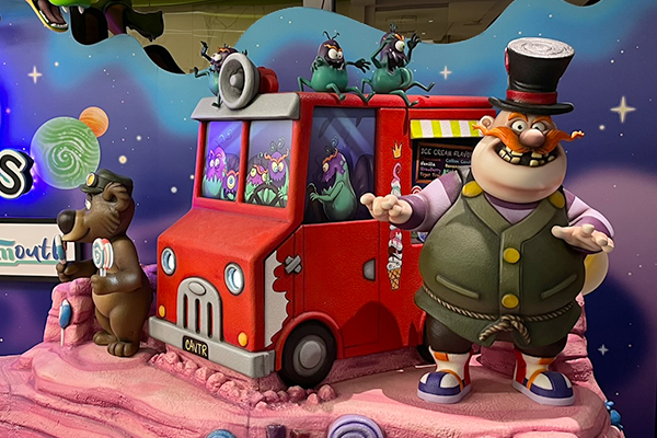 A colorful, whimsical display features a red ice cream truck with cartoon sugar bugs. Cavitar, mustached man in a top hat, and Molar Bear, a brown bear, stand beside the truck on a pink, fantasy-themed platform.