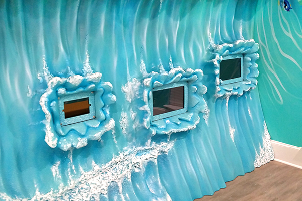 A sculpted wave wall features three rectangular video game framed by sculpted, foamy water, creating an underwater oceanic theme in a children's play area in a pediatric dental office.