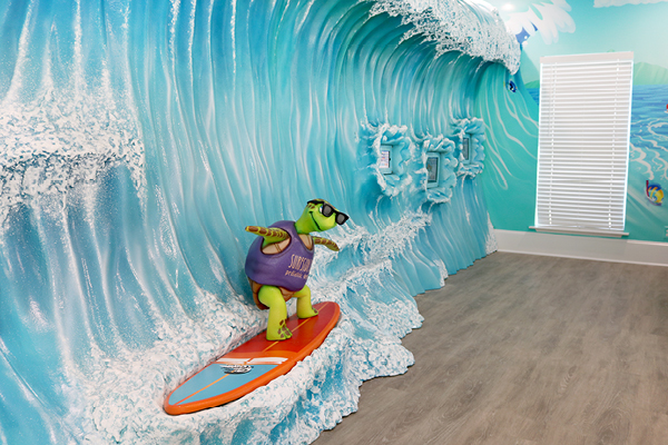 A colorful, playful game wall creates the perfect photo op with a large 3D wave mural, where a turtle statue wearing sunglasses and a purple vest surfs on an orange surfboard.