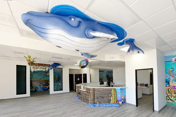 A reception area in a children’s dental office features large, blue whale sculptures hanging from the ceiling and an under-the-sea mural. A sign reads Kids Zone, and the decor is ocean-themed with bright, playful colors.