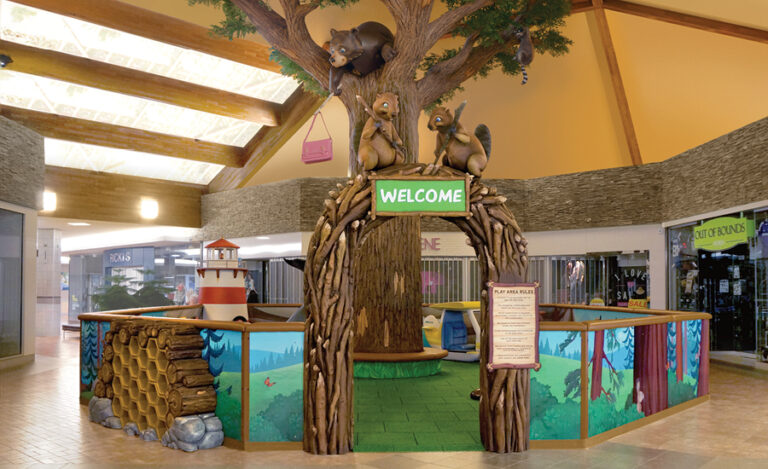 Indoor children’s mall play area themed like a forest, featuring a large tree with west coast animals and a “Welcome” sign at the entrance. The play space is surrounded by colorful forest-themed panels.
