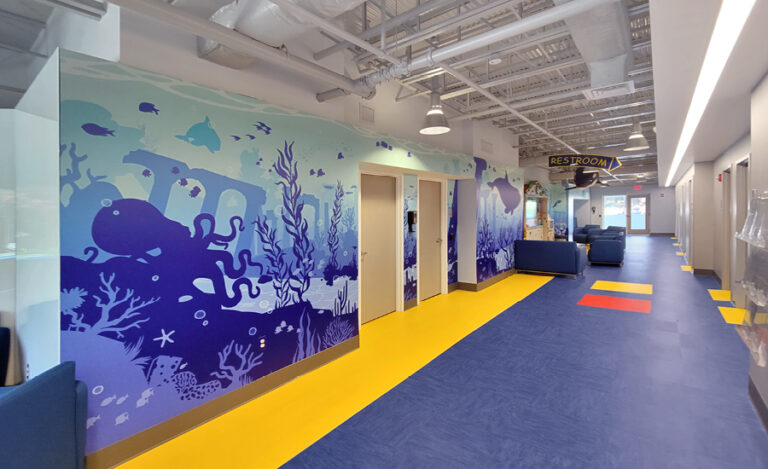 A bright healthcare facility hallway with a underwater-themed silhouette mural featuring an octopus, coral, and fish, with a complimentary blue and yellow floor, and blue seating.