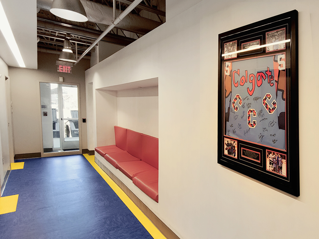 This is a boring before photo: a modern hallway with blue and yellow floor tiles, drab red cushioned bench seating built into a wall, and a framed Colgate sports jersey display. A glass door and exit sign are visible at the end.