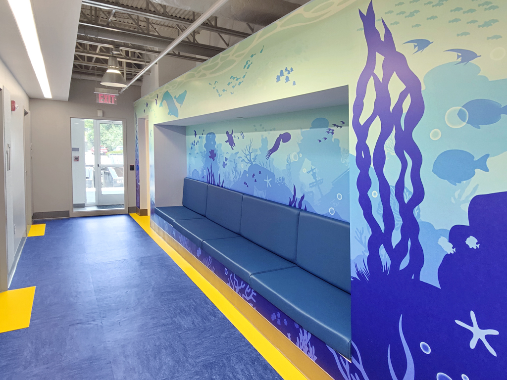 Experience a themed environment transformation: a hallway with blue and yellow flooring, a long blue bench, and walls adorned with an underwater ocean mural of fish, seaweed, and bubbles. Glass doors at the end lead outside.
