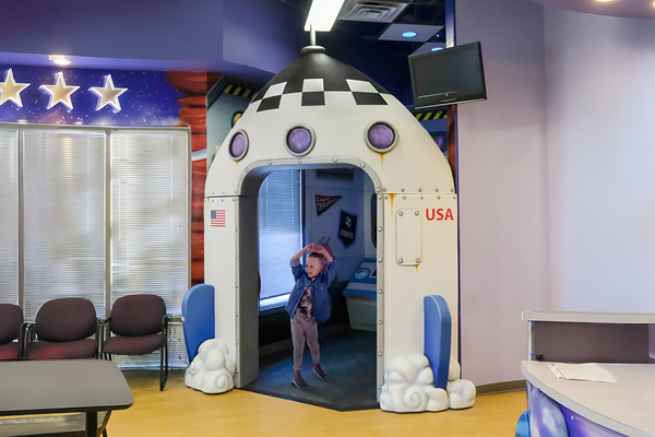 A young child stands under a rocket ship doorway in a colorful indoor room with space theme library decor, surrounded by chairs, a TV, and outer space-inspired decorations.