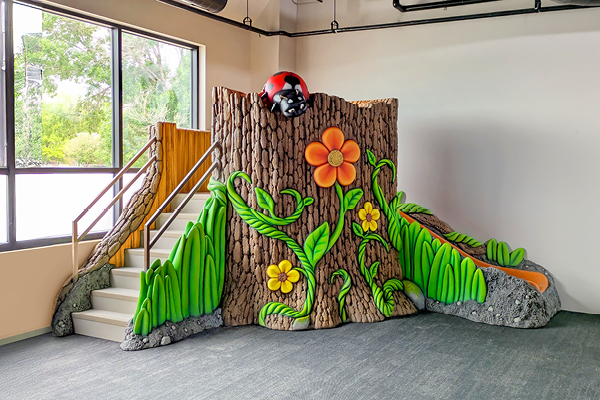 A colorful indoor playground slide shaped like a tree stump decorated with large flowers, green leaves, and a red ladybug on top, next to a staircase, in a bright room with large windows.