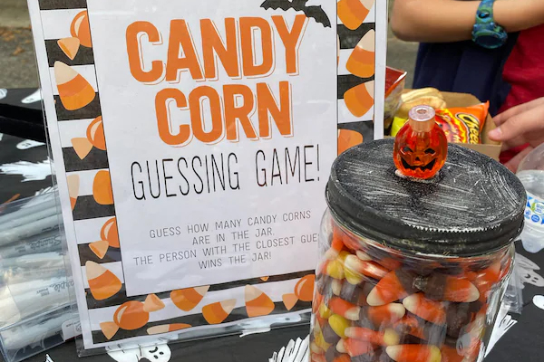 A sign reads Candy Corn Guessing Game! Guess how many candy corns are in the jar. The person with the closest guess wins the jar! Next to it is a jar filled with candy corn and topped with a jack-o’-lantern decoration.