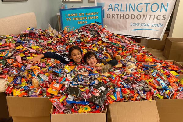 Two children smiling while lying in a massive pile of assorted candy, surrounded by donation boxes. A banner in the background reads ARLINGTON ORTHODONTICS and a sign says, I gave my candy to troops over seas.