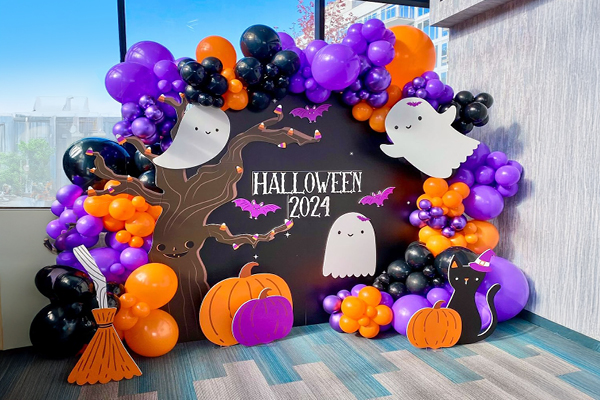 A Halloween 2024 photo booth with a black backdrop, cartoon ghosts, pumpkins, bats, a witchs broom, and a black cat, surrounded by clusters of orange, purple, and black balloons.