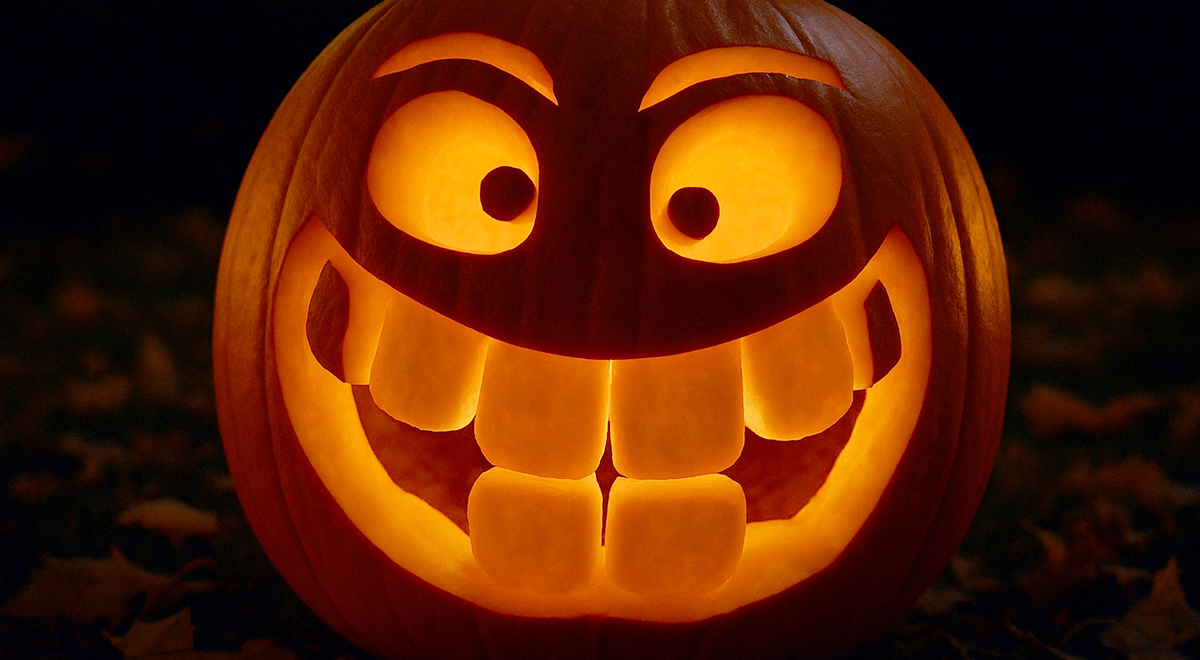 A glowing jack-o’-lantern with a mischievous, wide toothy grin and arched eyebrows carved into its face, illuminated from within against a dark background.