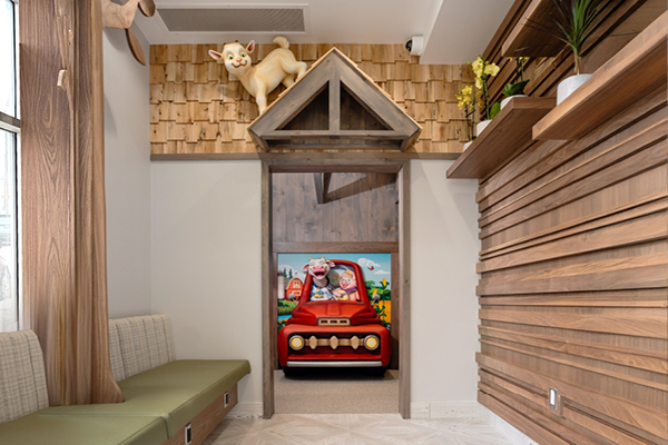 A playful waiting room with wooden walls, green cushioned benches, a cat statue above a door, and a red toy car with cartoon animals in a colorful play area beyond the doorway.