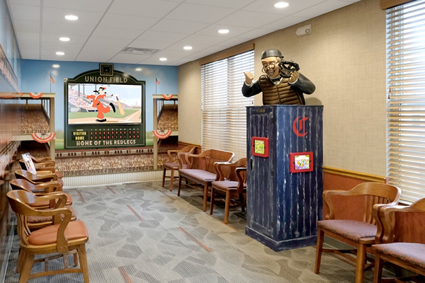 A waiting room decorated with a baseball theme, featuring wooden chairs, a mural of a stadium scoreboard, and a statue of an umpire in full gear standing on a blue platform. Light streams in from tall windows with blinds.