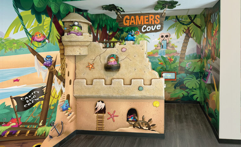 A colorful playroom with a pirate and beach theme, featuring a sandcastle structure, cartoon pirates, and tropical mural walls. The sign above the castle reads GAMERS Cove..