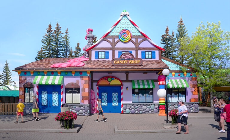 A whimsical, colorful candy shop with a gingerbread house design, bright blue doors, candy decorations, and striped awnings. People walk nearby on a sunny day with trees in the background.