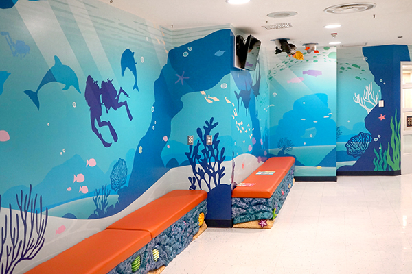 A waiting area with orange benches and walls decorated with a colorful underwater mural featuring dolphins, fish, coral, seaweed, and a scuba diver. The floor is white and the space is brightly lit.