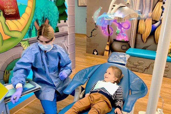 A dentist in blue scrubs and a mask prepares tools while a smiling child lies in a dental chair. The colorful, fantasy-themed office features dragons, castle walls, and floating magical elements on the wall.
