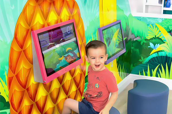 A young boy with short hair and a red shirt smiles while sitting in front of a touchscreen display in a colorful, jungle-themed play area with leafy wall art.