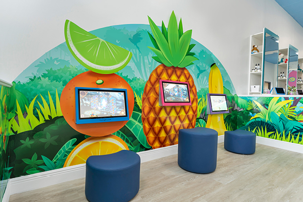 A colorful kids’ area with blue stools, wall-mounted tablets, and a vibrant mural featuring large fruit illustrations, including an orange, lime, and pineapple, set against a jungle-themed background.