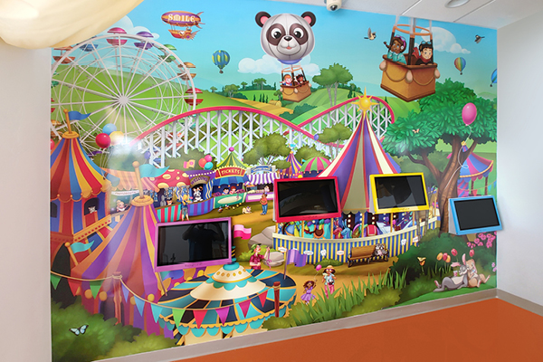 A colorful mural of a cartoon amusement park with a ferris wheel, roller coaster, circus tents, and a panda in a cable car. Four black screens are mounted on the wall in front of the mural.