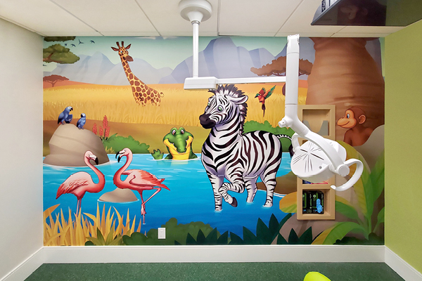 A colorful mural of a savanna scene in a children’s room, featuring a zebra, giraffe, flamingos by a pond, crocodile, birds, and a monkey. Dental equipment hangs from the ceiling in the foreground.