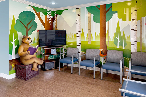 Colorful waiting room with a forest-themed mural, gray chairs, a TV, and a bear statue holding a book, creating a playful and welcoming atmosphere for children.