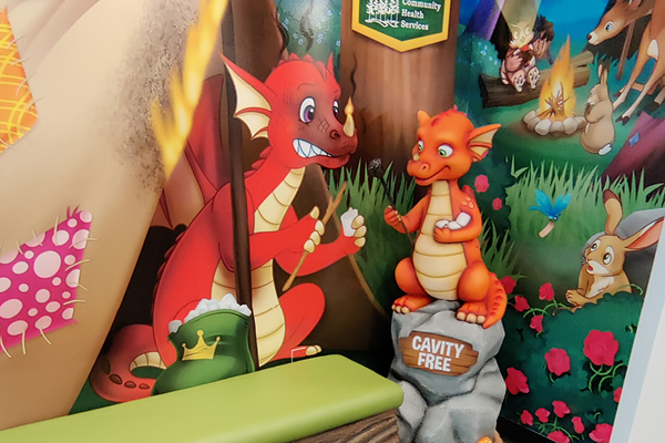 A mural shows a large red dragon helping a smaller orange dragon, who sits on a rock labeled CAVITY FREE. The background includes cartoon animals, colorful plants, and a campfire scene.
