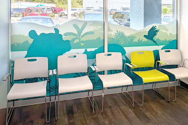 A row of white chairs with one bright yellow chair in a waiting area; behind them is a mural with silhouettes of animals, plants, and a bird, and large windows showing parked cars outside.