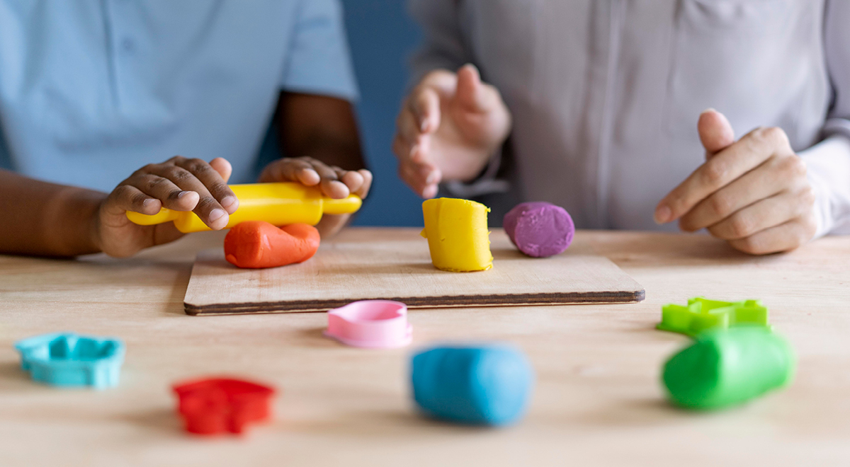 Two people at a table use colorful modeling clay and cookie cutters. One person rolls yellow clay with a small roller while various clay shapes and tools are scattered on the table.