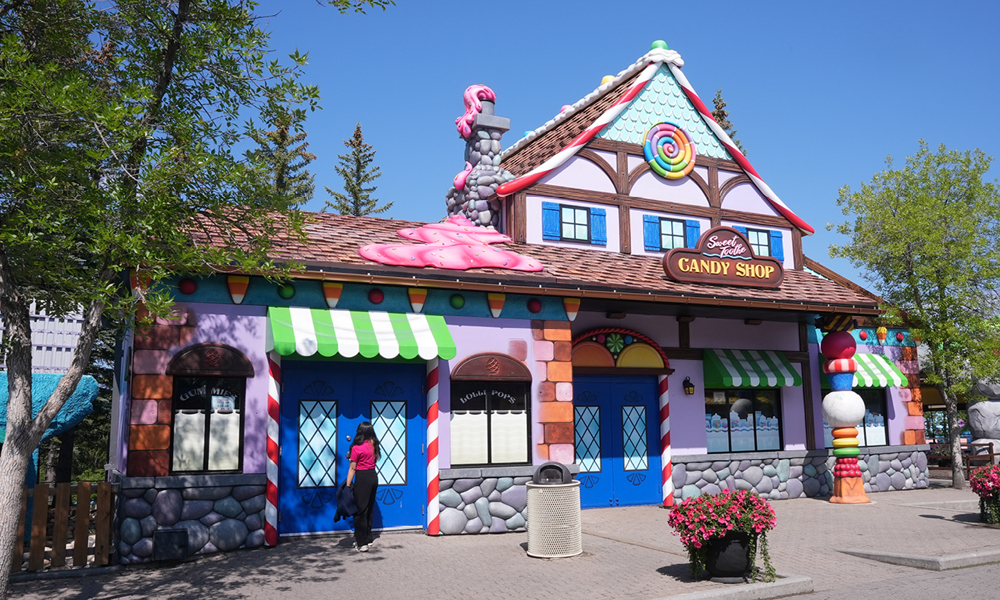 A whimsical candy shop with colorful decorations, striped awnings, candy-shaped ornaments, and a roof topped with pink icing. A person in pink stands near the blue doors beneath a "Now Hiring" sign as trees frame the scene.