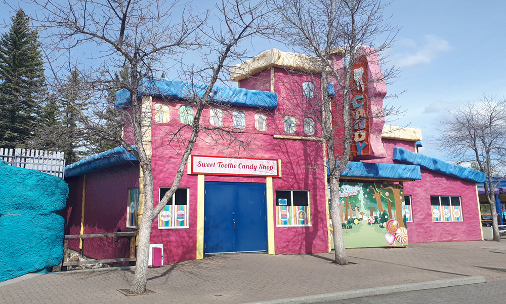 A brightly colored candy shop with pink walls, blue trim, and a large “CANDY” sign. Leafless trees stand in front, colorful posters decorate the windows, and a “Now Hiring” sign hangs beside the door of Sweet Tooth Candy Shop.
