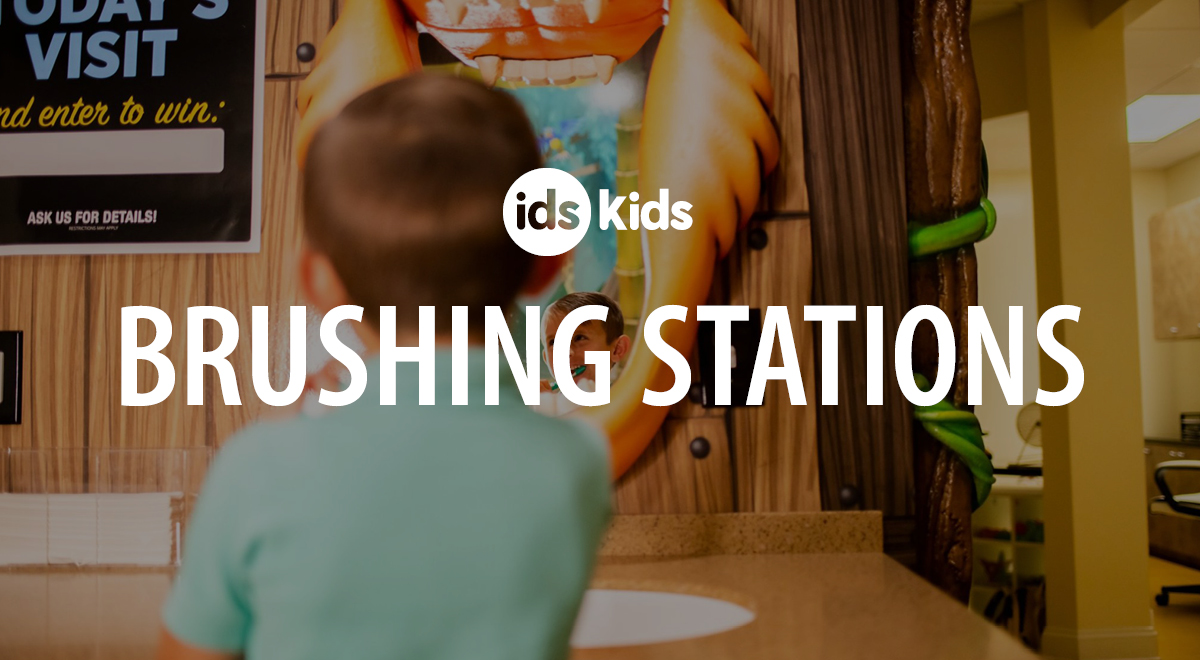 A young child stands at a sink in front of a wall sign that reads ids Kids Brushing Stations in a brightly lit, kid-friendly dental office.
