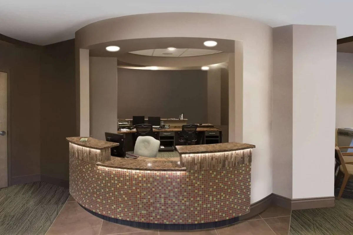 A modern, curved reception desk with colorful mosaic tiles, brown countertops, and a white chair sits in front of a workspace with computers and office chairs. The area is well-lit with neutral-colored walls—this is a drab before photo.