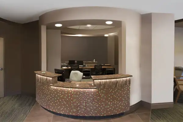 A modern, curved reception desk with colorful mosaic tiles, brown countertops, and a white chair sits in front of a workspace with computers and office chairs. The area is well-lit with neutral-colored walls—this is a drab before photo.