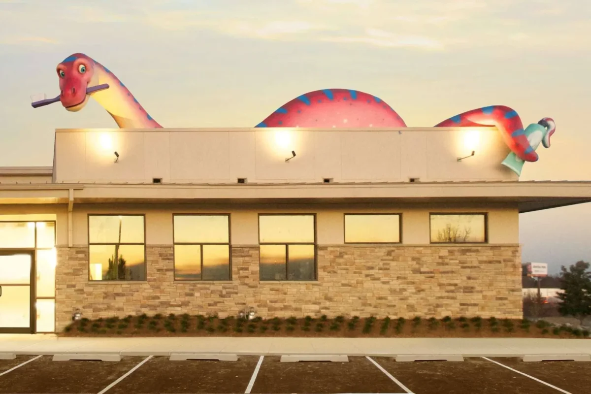 Before and after a themed environment transformation, a large, colorful sculpture of a smiling dinosaur holding a toothbrush now sits atop the tan brick dental office, glowing warmly in the sunset.