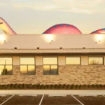 Before and after a themed environment transformation, a large, colorful sculpture of a smiling dinosaur holding a toothbrush now sits atop the tan brick dental office, glowing warmly in the sunset.