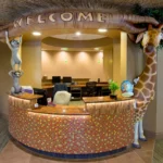 Experience a themed environment transformation: A colorful reception desk with a mosaic tile front, decorated with a large WELCOME sign held by playful animal statues, including a giraffe and lemurs, in a cheerful, jungle-themed office lobby.