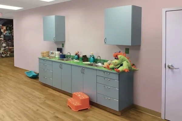 A clinic exam room with light blue cabinets, a green countertop, and a toy dinosaur on the counter shows anything but a boring before photo—colorful toy sinks, cups, and wooden flooring brighten the space, while a closed door sits to the right.