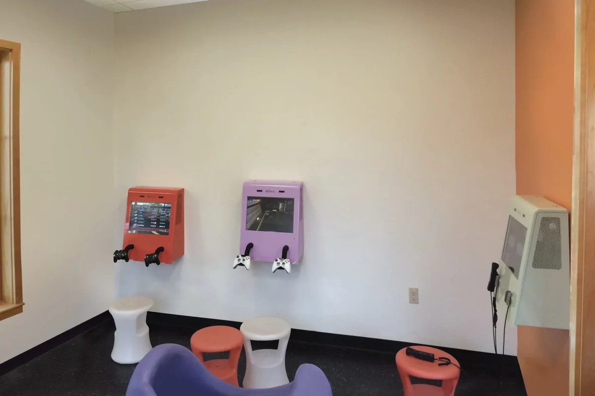 This is a boring before photo: a small room with drab pastel-colored walls, two wall-mounted gaming stations with attached controllers, mismatched stools, and an additional wall device with a telephone-style handset.