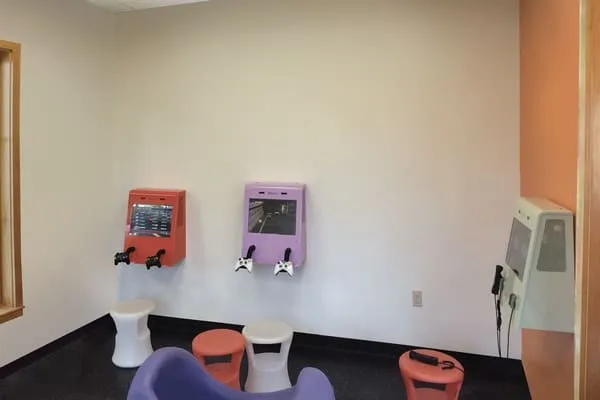 This is a boring before photo: a small room with drab pastel-colored walls, two wall-mounted gaming stations with attached controllers, mismatched stools, and an additional wall device with a telephone-style handset.