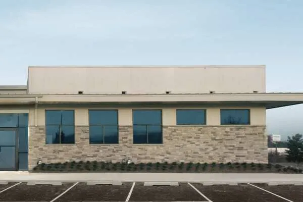 A modern commercial building with large blue-tinted windows, drab stone and stucco exterior, flat roof, and an empty parking lot in front under a clear sky—this is a boring before photo.
