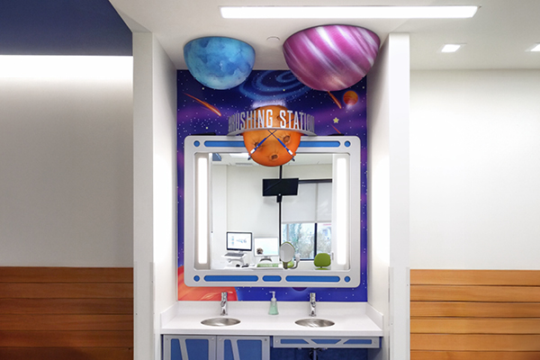 A colorful handwashing station with a space theme, featuring painted planets on the ceiling, a mirror framed like a spaceship window, and two sinks below. The wall has stars, planets, and the words “WASHING STATION.”.