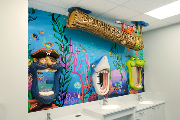 A colorful underwater-themed kids’ bathroom wall featuring 3D cartoon heads—a pirate, a shark, and an alligator—with wide mouths above sinks, and a sign reading “Brushing Station” on a wooden log.