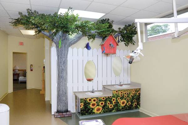 A dental office corner decorated with a tree, white picket fence, birdhouse, and sunflowers. Two oval mirrors are set above sunflower-themed counters, with dental equipment visible on the right.