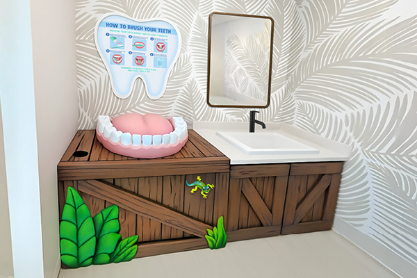 A childrens dental hygiene station with a large model of teeth, a tooth-shaped sign showing brushing instructions, a sink with a mirror, and wooden-themed cabinets decorated with green plants and a lizard.