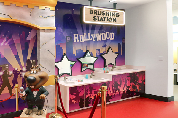 A Brushing Station with a Hollywood theme, featuring star-shaped mirrors, a cartoon bear statue in a tuxedo holding a microphone, and a vibrant backdrop of city silhouettes and cameras. Red rope barriers are in front.