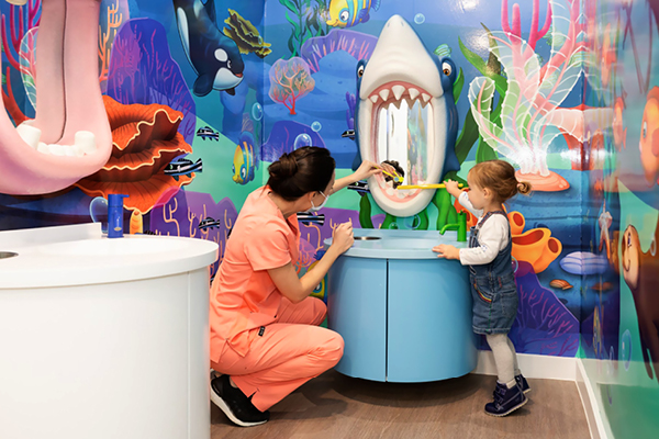 A dental hygienist in peach scrubs helps a young girl brush a toy crocodiles teeth using a large shark display in a colorful, ocean-themed childrens dental office.
